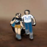The Couple enamel pin—a couple walking down the street and talking, as illustrated by Jiaqi Wang— shown resting on a wood block.