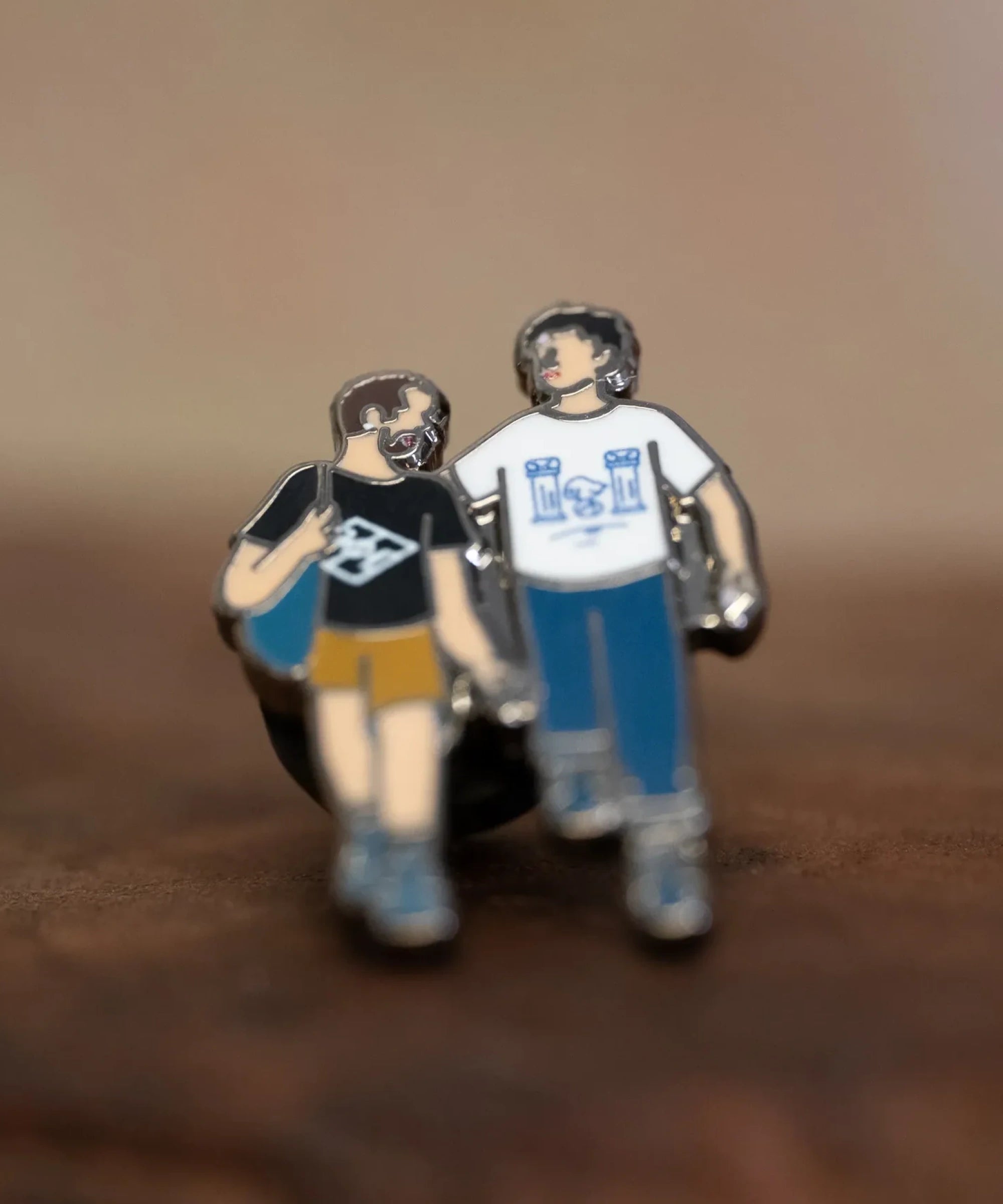 The Couple enamel pin—a couple walking down the street and talking, as illustrated by Jiaqi Wang— shown resting on a wood block.