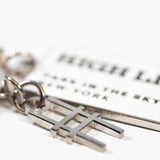 Closeup of the High Line Tag Keychain, featuring a small charm of the High Line railroad-track "H" icon in focus, with the enamel tag out of focus.