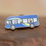 The MTA Bus enamel pin—MTA Bus with driver and passengers, including the sightseer, illustrated by Jiaqi Wang— resting on a wood block.