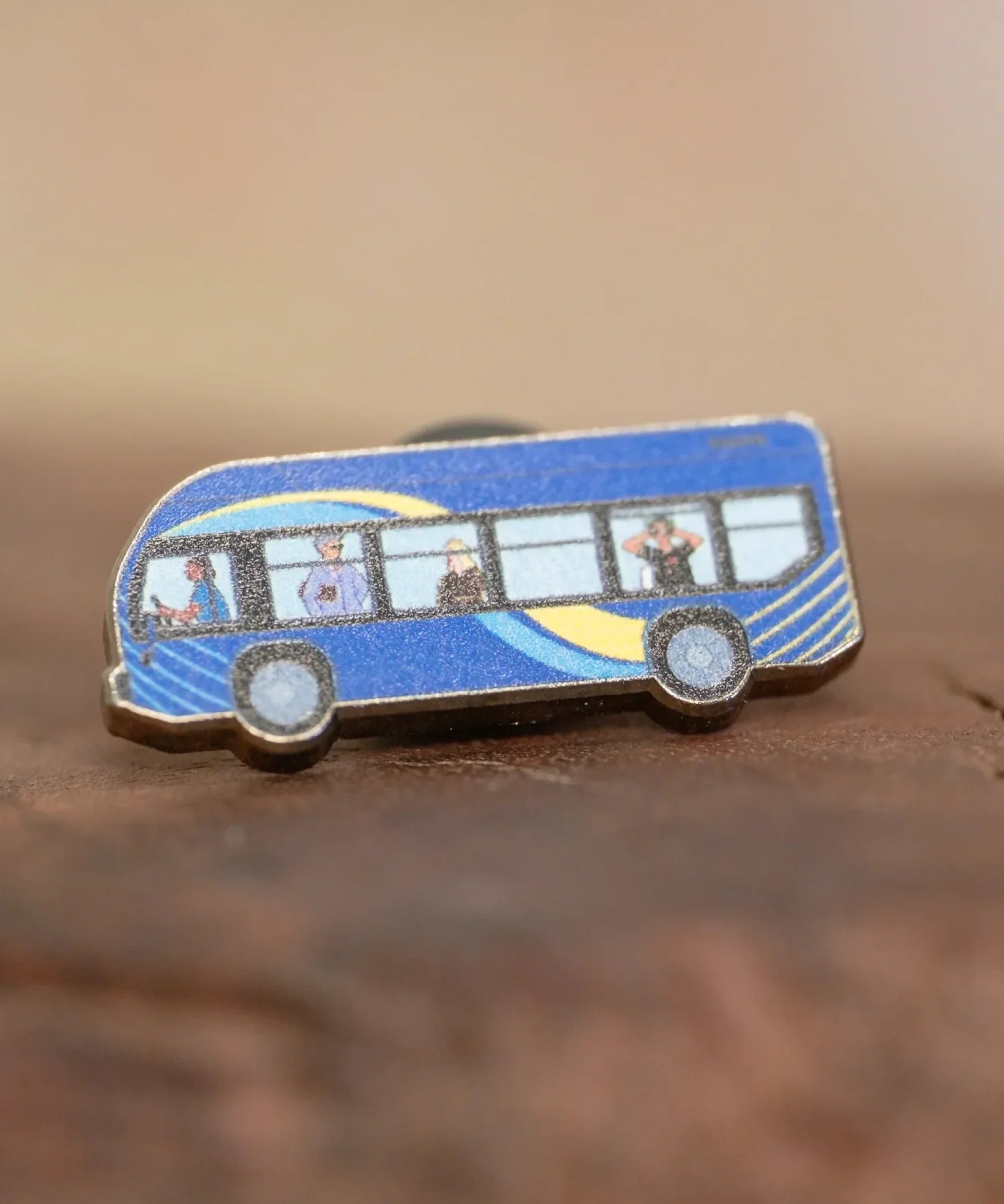 The MTA Bus enamel pin—MTA Bus with driver and passengers, including the sightseer, illustrated by Jiaqi Wang— resting on a wood block.