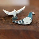The Pigeon Pins—two enamel pigeon pins, one sitting and one in flight—resting on a wood block.