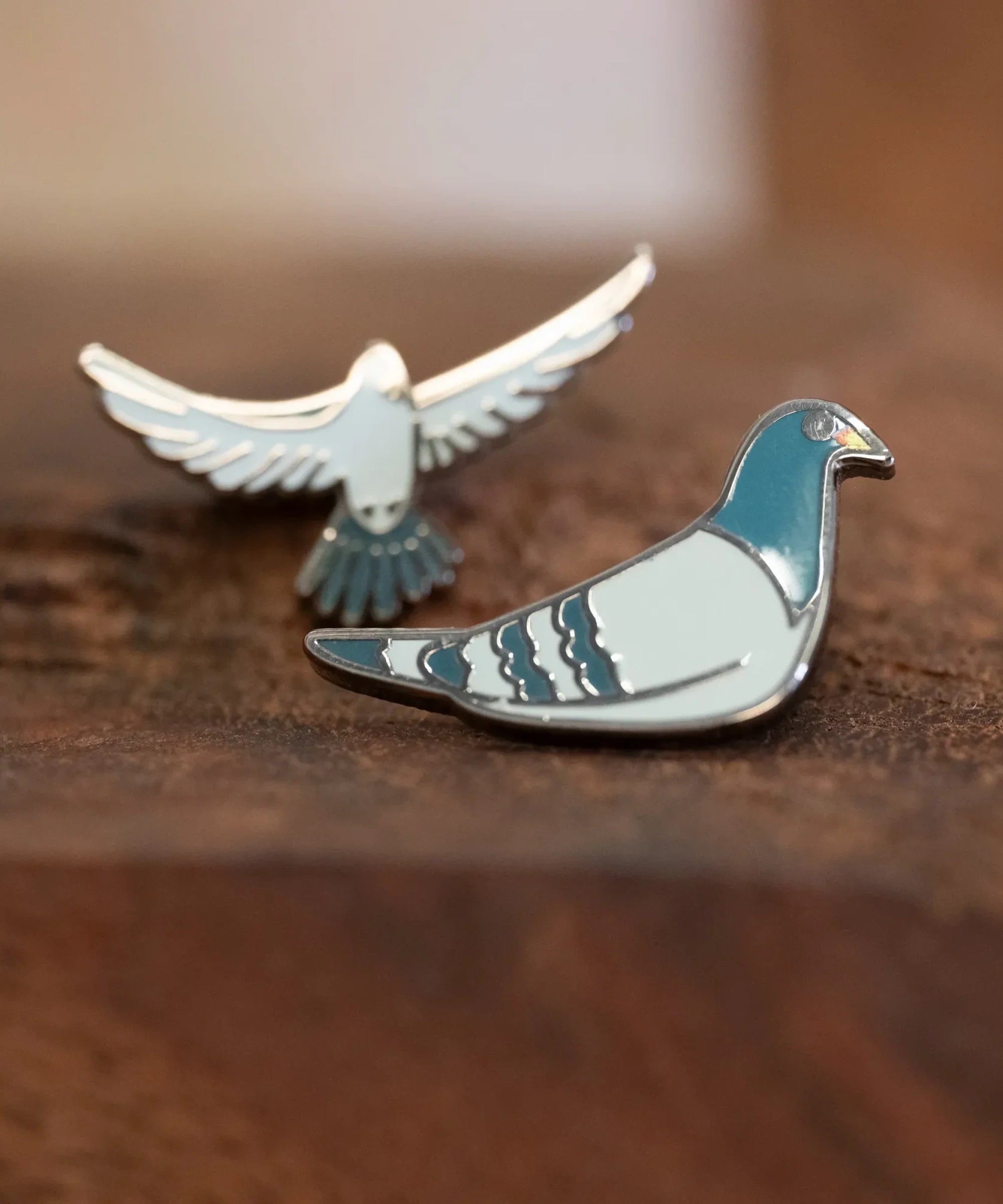 The Pigeon Pins—two enamel pigeon pins, one sitting and one in flight—resting on a wood block.