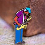 Saxaphone Player enamel pin—a street musician playing the saxophone as illustrated by Jiaqi Wang. The pin leans against a wood block.
