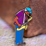 Saxaphone Player enamel pin—a street musician playing the saxophone as illustrated by Jiaqi Wang. The pin leans against a wood block.