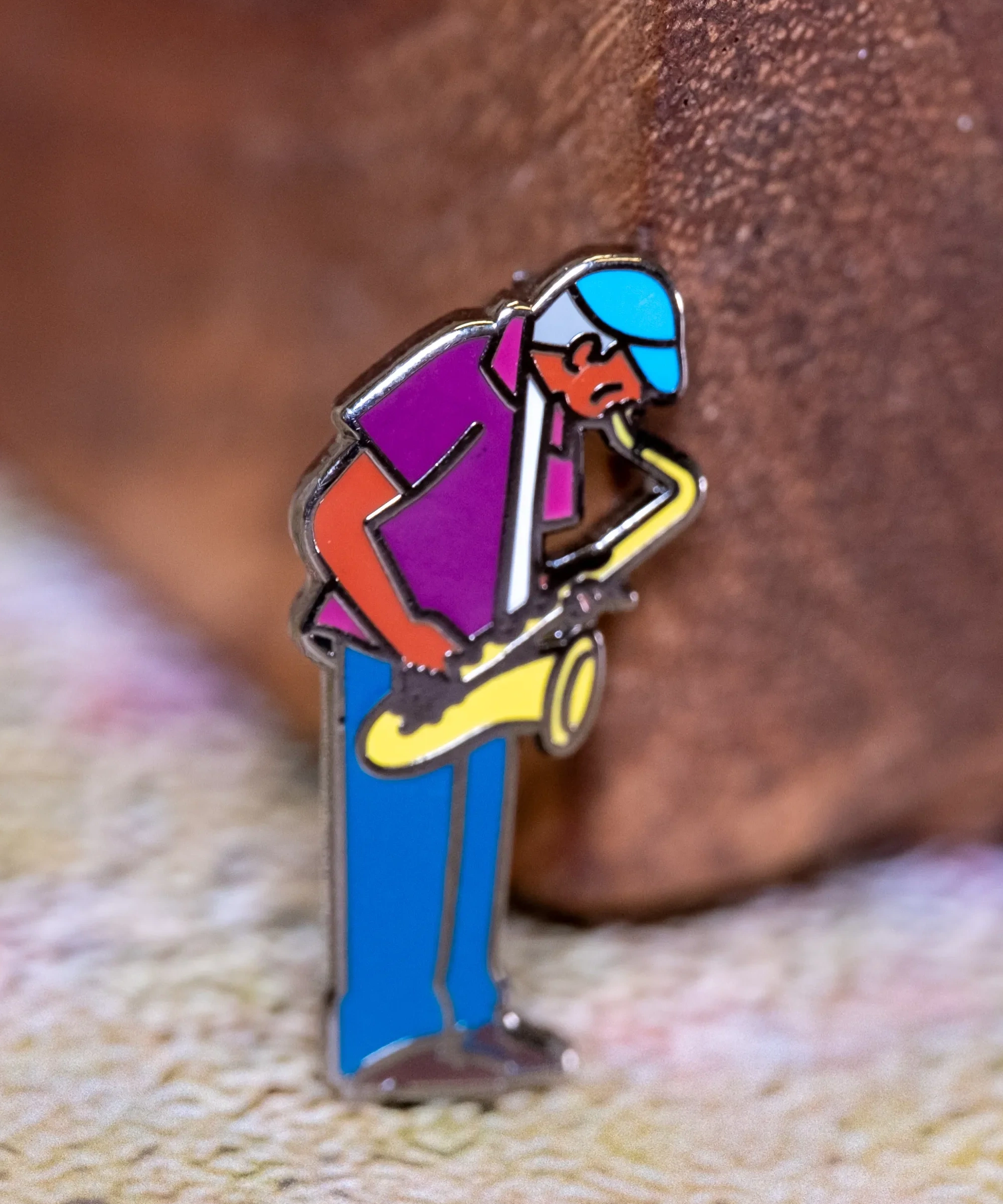 Saxaphone Player enamel pin—a street musician playing the saxophone as illustrated by Jiaqi Wang. The pin leans against a wood block.