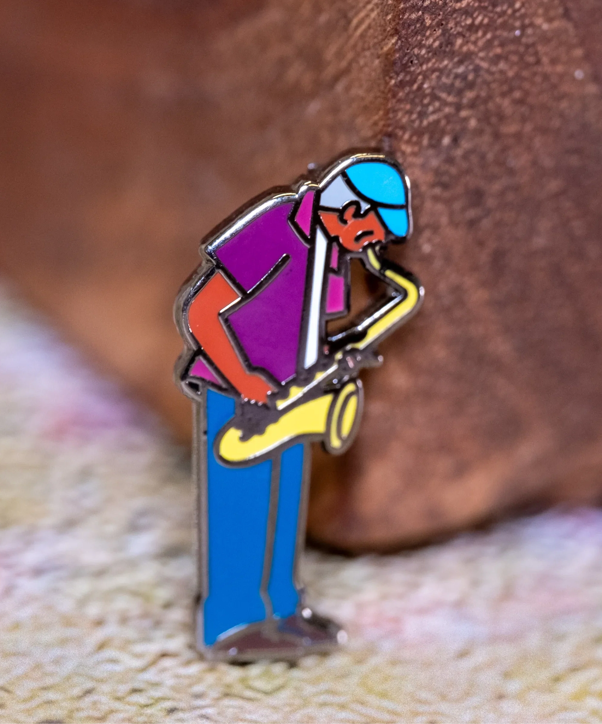 Saxaphone Player enamel pin—a street musician playing the saxophone as illustrated by Jiaqi Wang. The pin leans against a wood block.