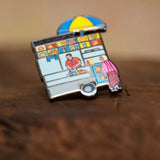 Street Vendor enamel pin—NYC street food cart—resting against a wood block.