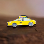 Taxi enamel pin—a NYC classic yellow cab illustrated by Jiaqi Wang—resting on a wood block.