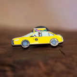 Taxi enamel pin—a NYC classic yellow cab illustrated by Jiaqi Wang—resting on a wood block.