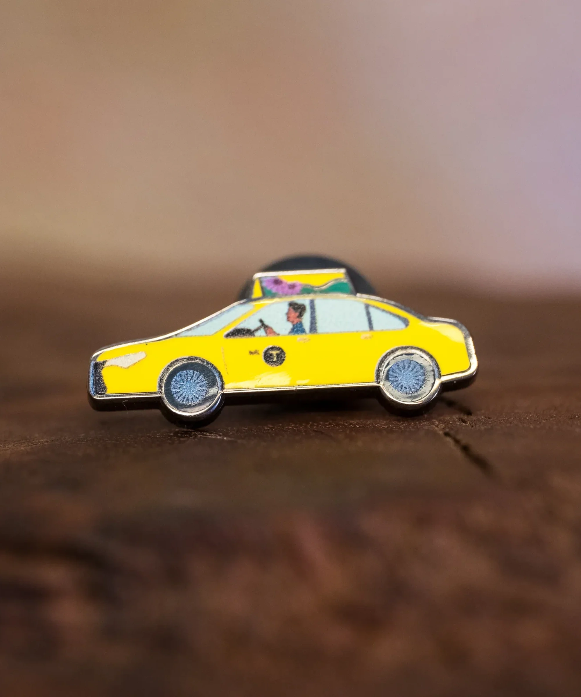 Taxi enamel pin—a NYC classic yellow cab illustrated by Jiaqi Wang—resting on a wood block.