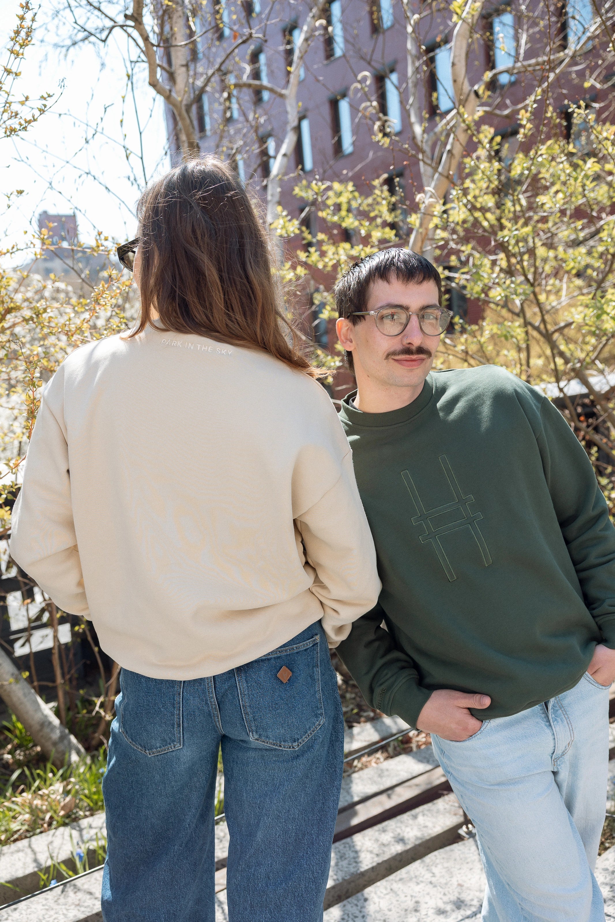 Two people, each wearing the Big Icon Crewneck and standing on the High Line. The person wearing the Ivy sweatshirt looks at the camera, hands in their pockets.
