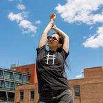 Woman wearing the High Line Icon Tee in black and stretching, the view of the Meatpacking district in the background.