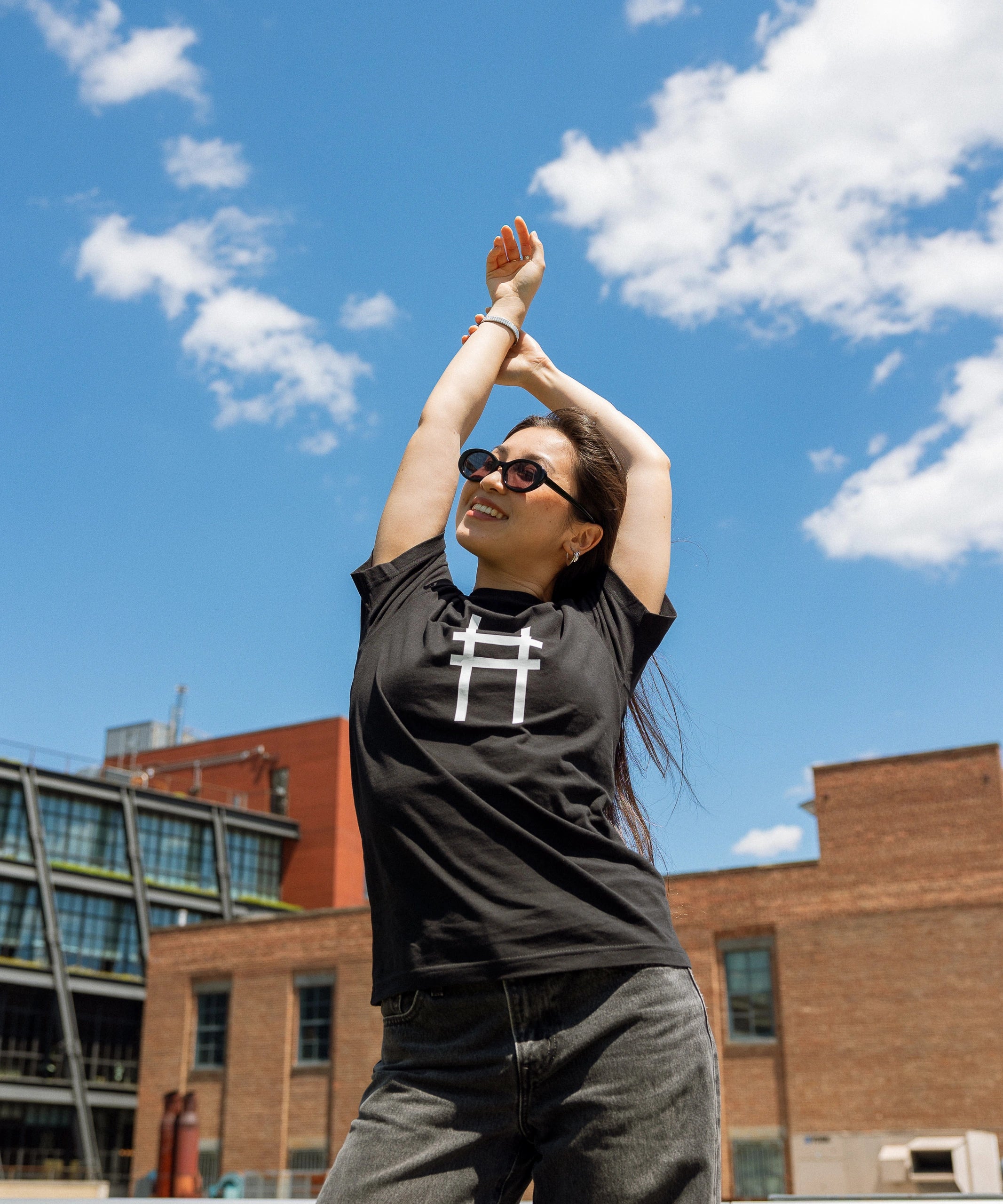 Woman wearing the High Line Icon Tee in black and stretching, the view of the Meatpacking district in the background.