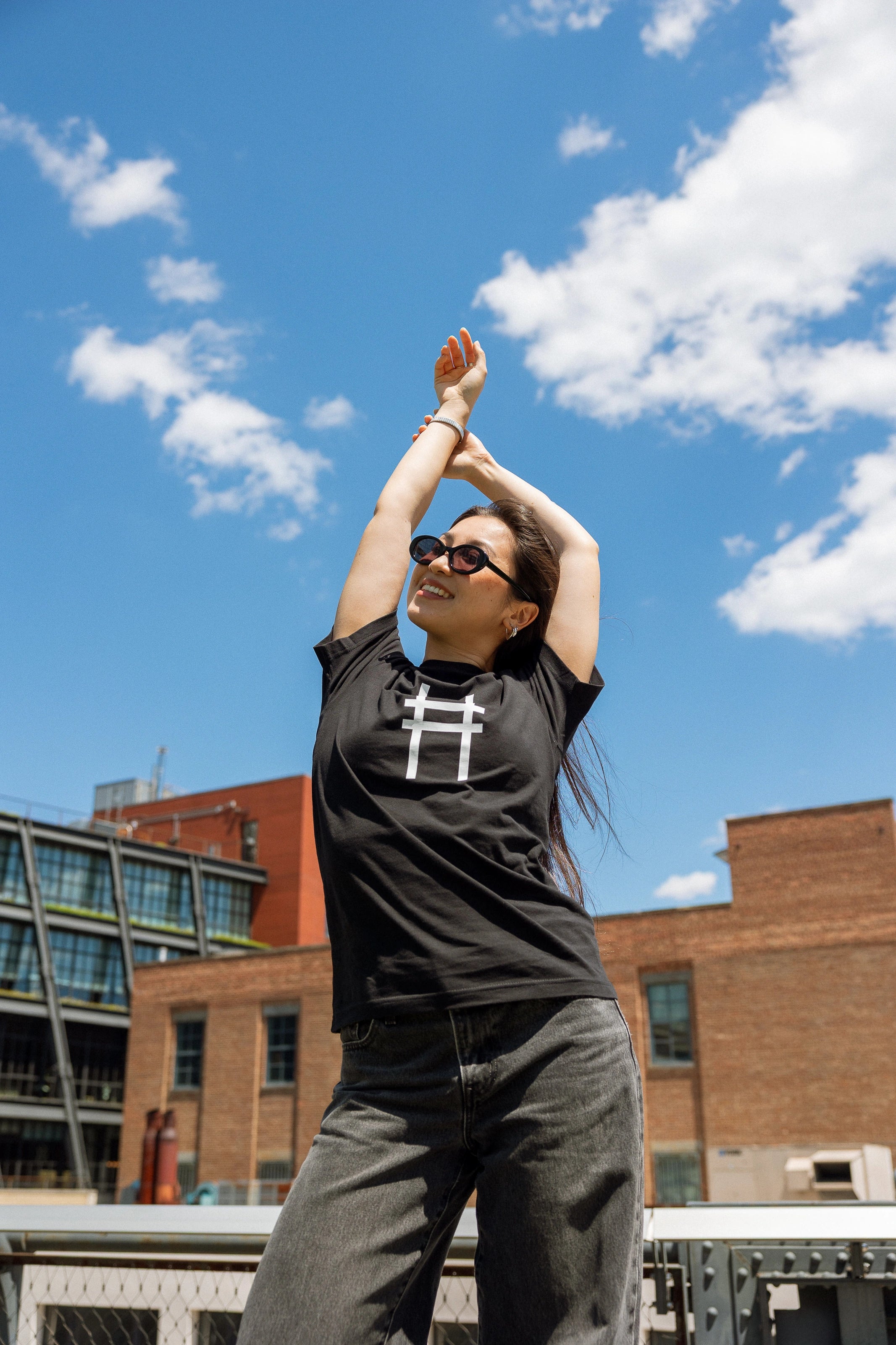 Woman wearing the High Line Icon Tee in black and stretching, the view of the Meatpacking district in the background.