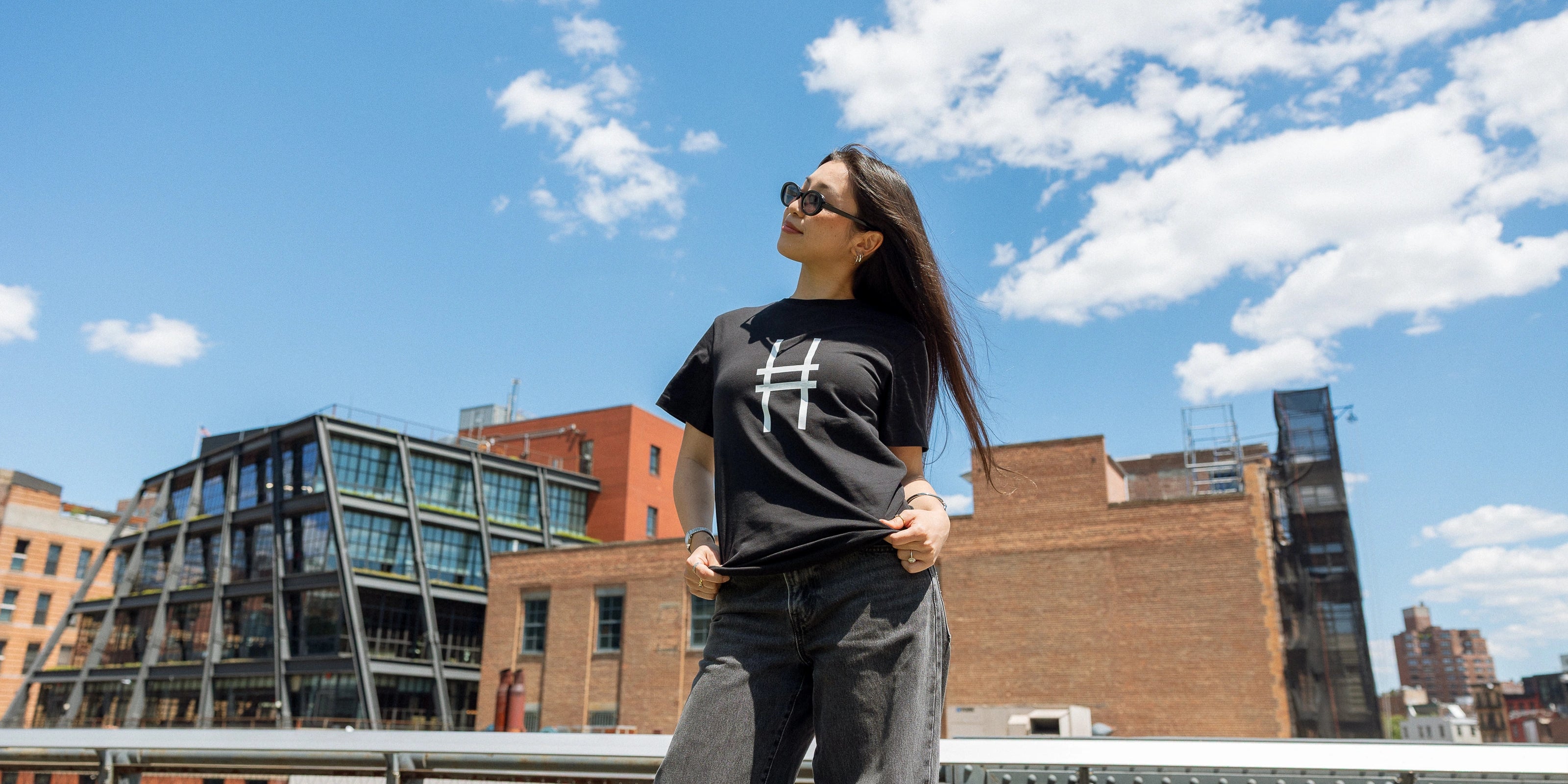 Woman wearing the High Line Icon Tee in black against the blue sky and view from the High Line.