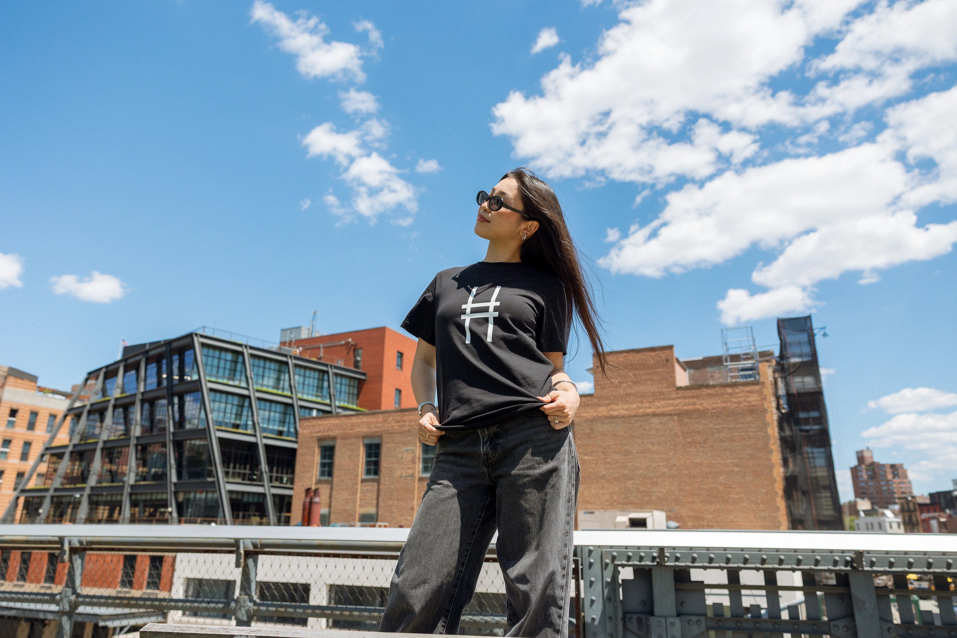 Woman wearing the High Line Icon Tee in black against the blue sky and view from the High Line.