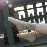 The Lingua Franca x High Line 100% Cashmere Socks on someone's feet against the historic railing of the High Line. These oatmeal colored socks are embroidered with the High Line icon in green just below the outer cuff of each sock.