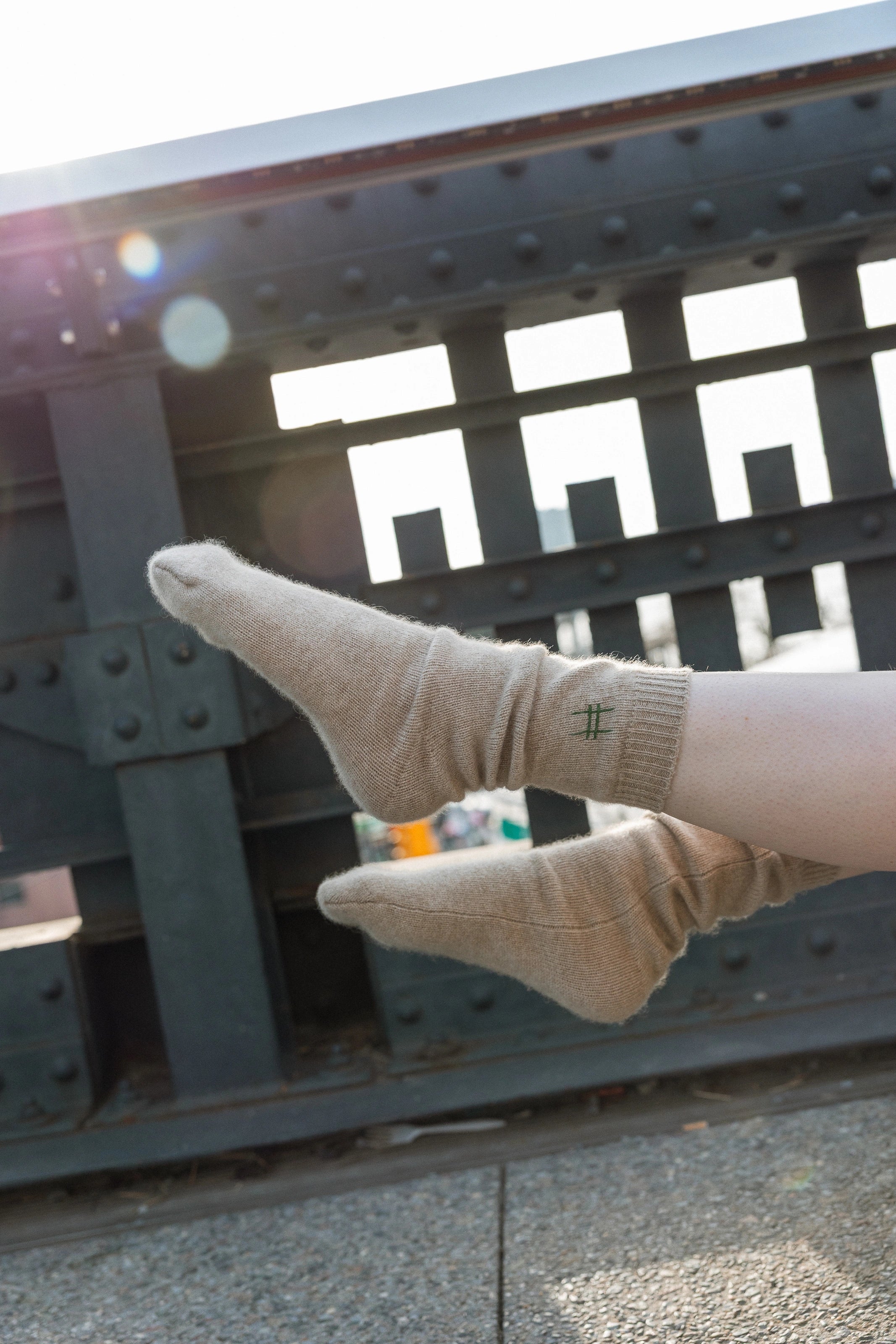The Lingua Franca x High Line 100% Cashmere Socks on someone's feet against the historic railing of the High Line. These oatmeal colored socks are embroidered with the High Line icon in green just below the outer cuff of each sock.