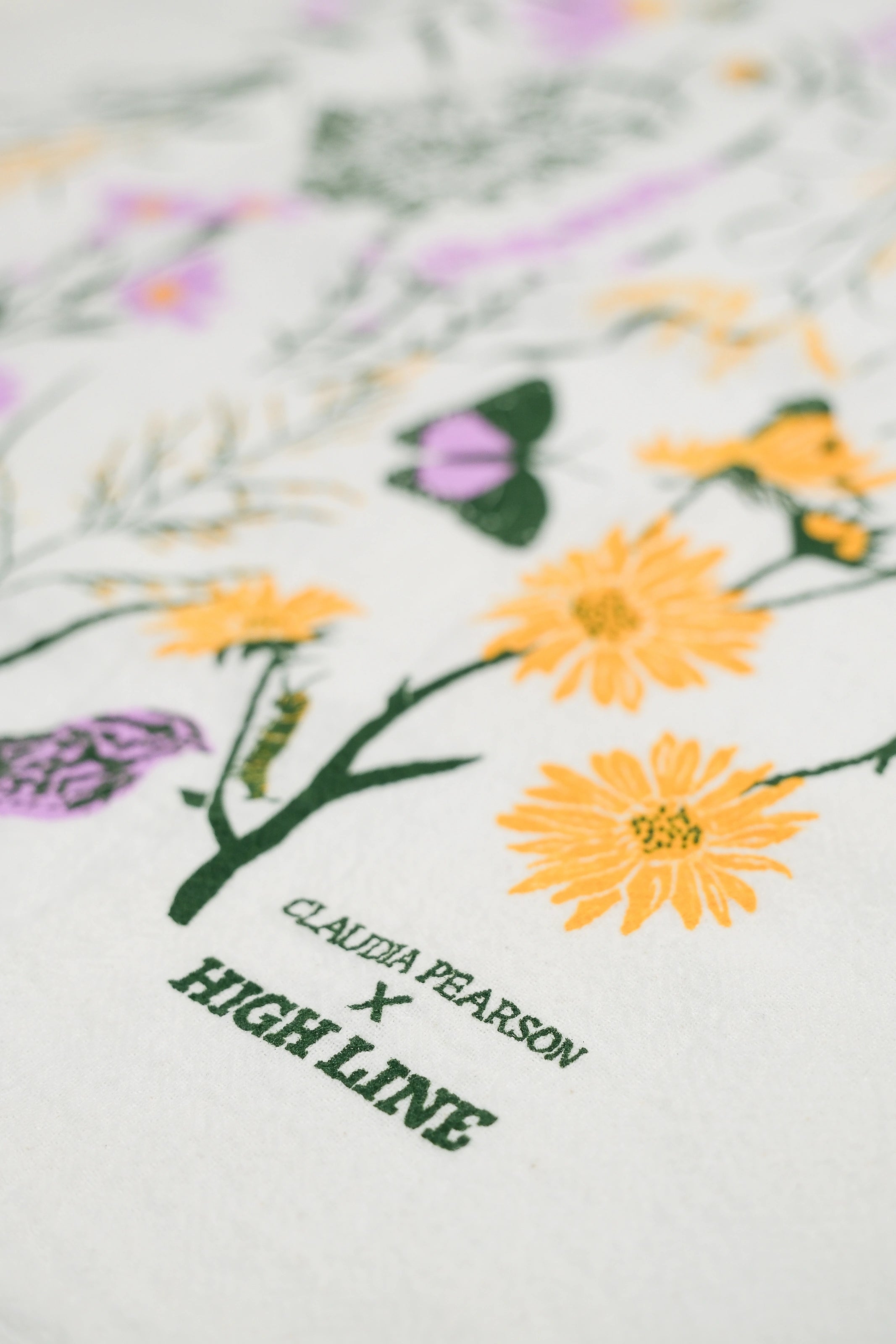 Claudia Pearson x High Line Chelsea Grassland tea towel in closeup. The flour sack cotton tea towel is covered in illustrations of the flowers and grasses that grow in the Chelsea Grassland area of the High Line in pale purple, deep green and yellow.