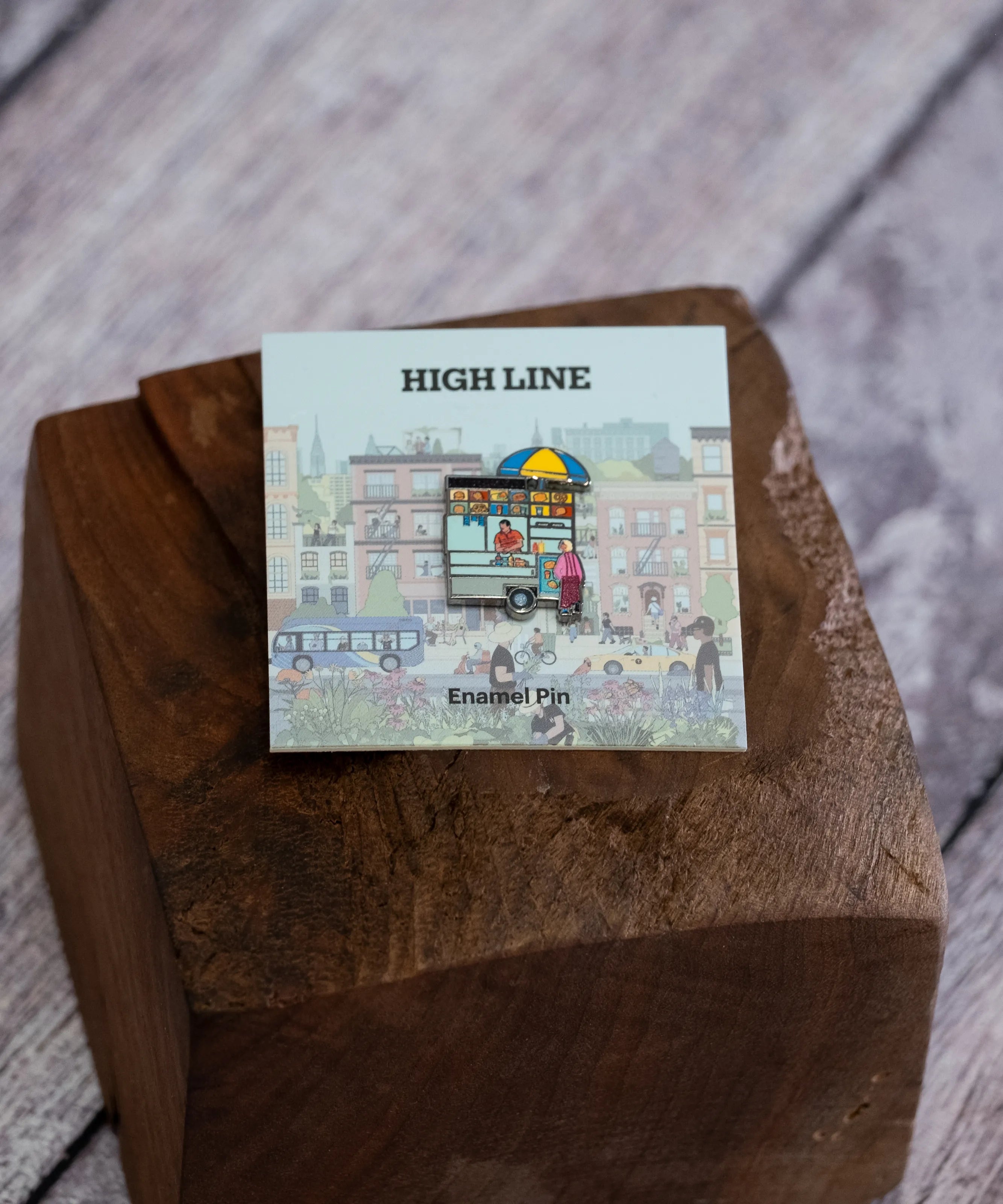 Street Vendor enamel pin—NYC street food cart—shown against the backing card and resting on a wood block.
