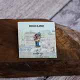 Saxaphone Player enamel pin—a street musician playing the saxophone as illustrated by Jiaqi Wang— shown on the backing card resting on a wooden block.