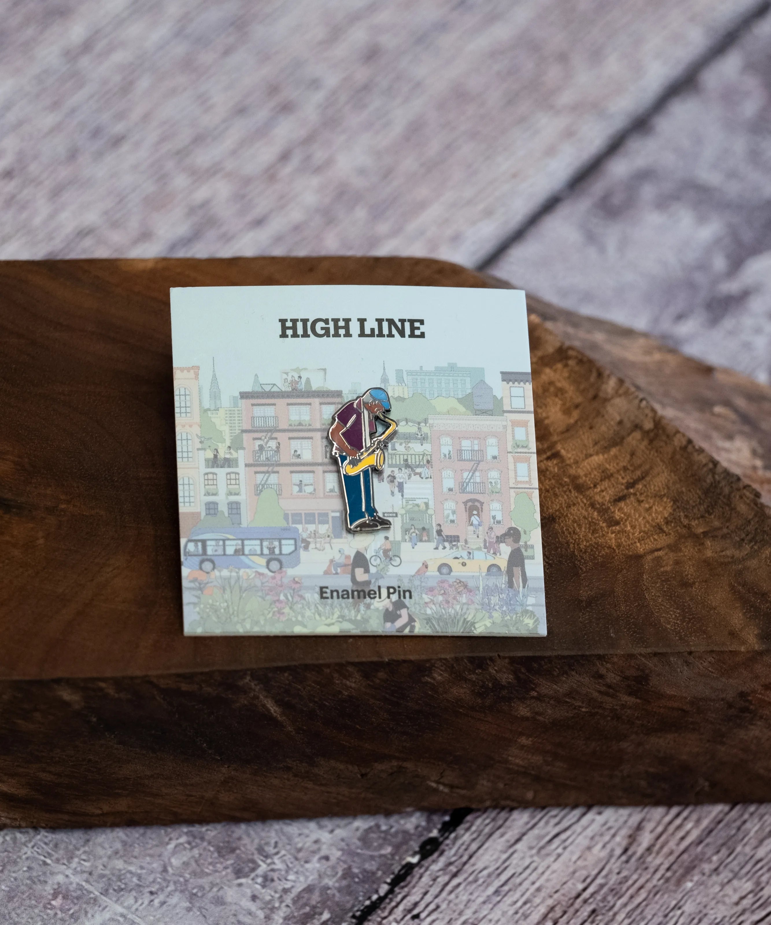 Saxaphone Player enamel pin—a street musician playing the saxophone as illustrated by Jiaqi Wang— shown on the backing card resting on a wooden block.