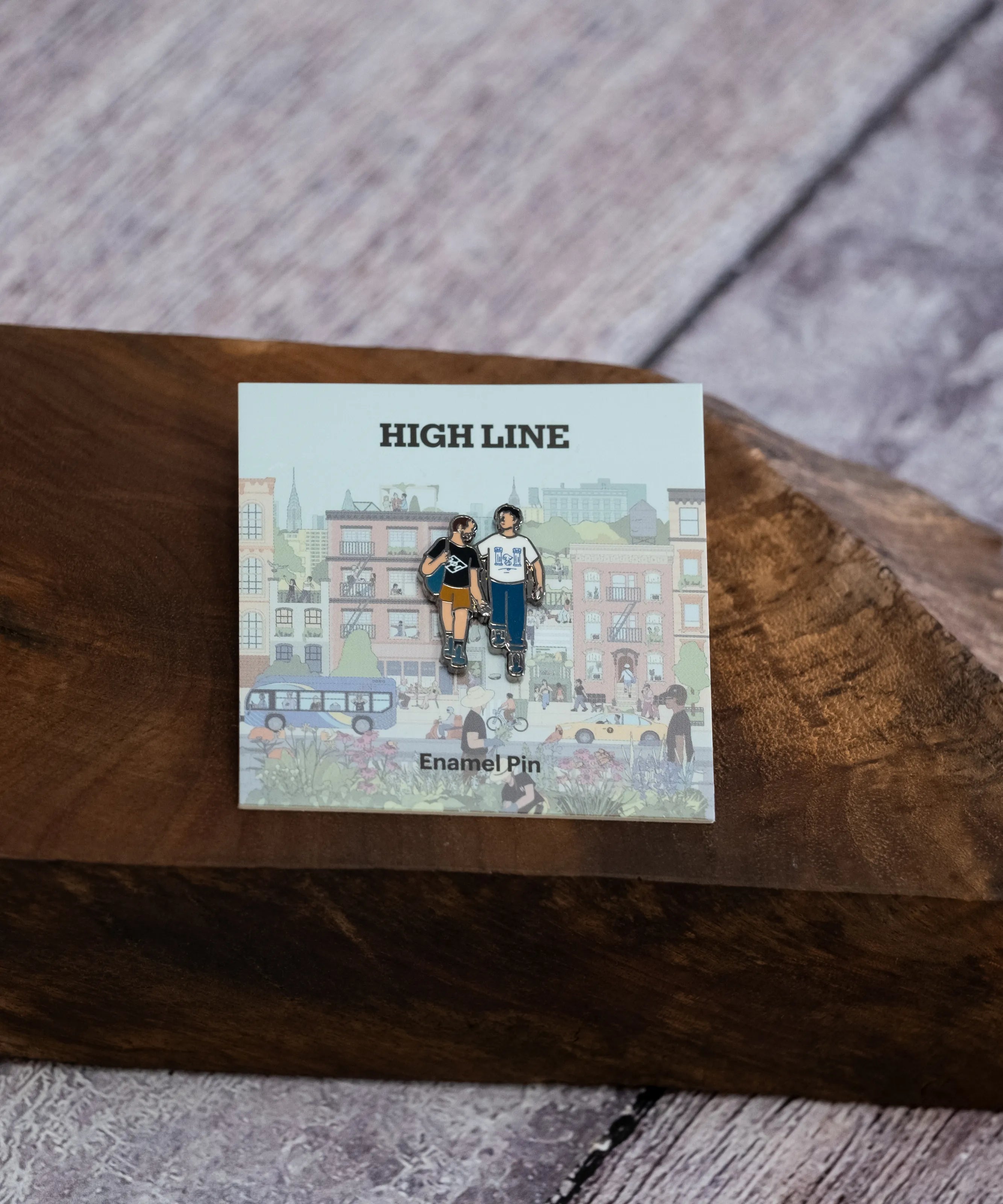 The Couple enamel pin—a couple walking down the street and talking, as illustrated by Jiaqi Wang— shown against the backing card and resting on a wood block.