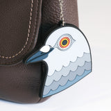 A close-up of a brown textured leather bag with the Dinosaur bag charm on it. The charm is a pigeon head inspired by Iván Argote's Dinosaur.