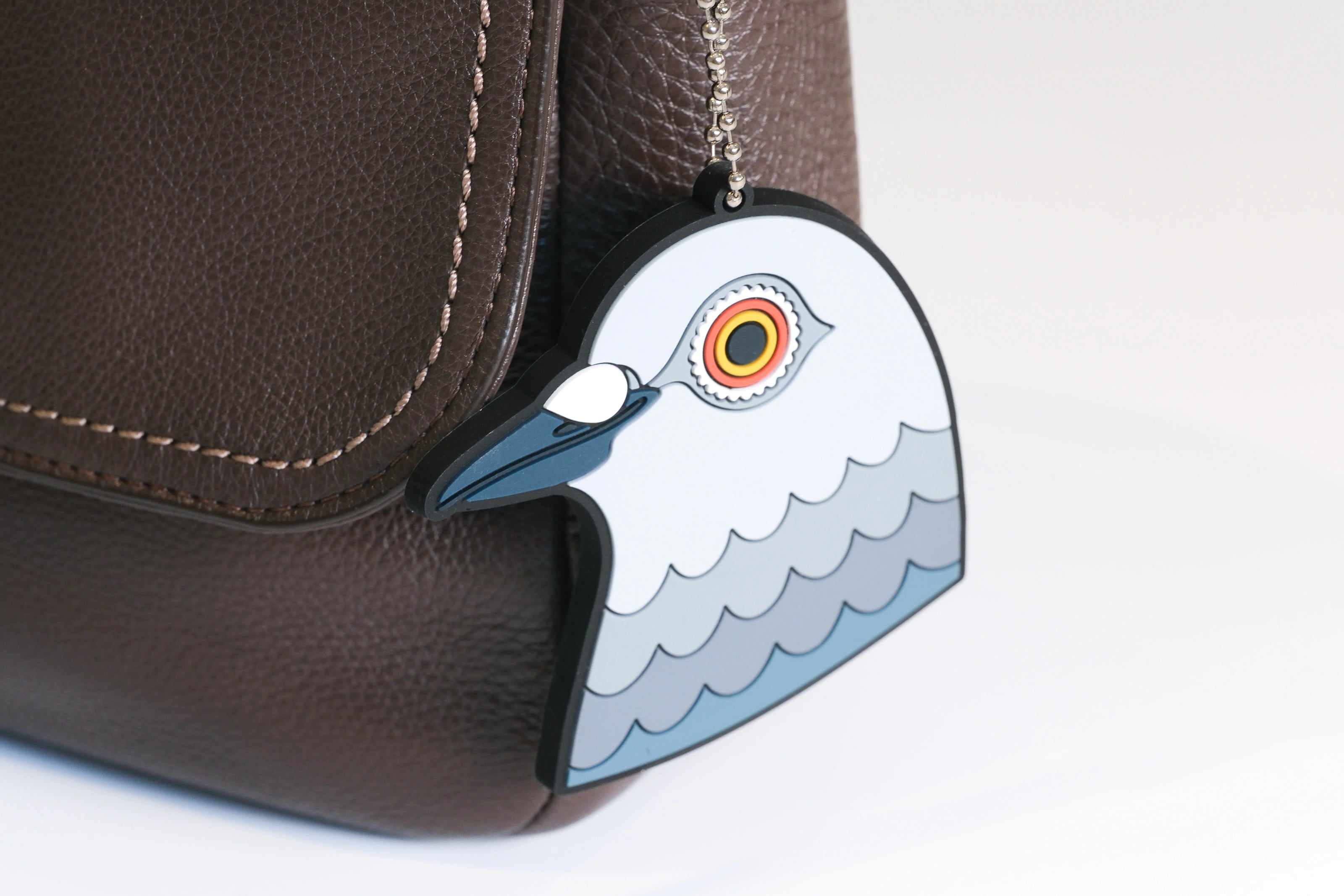 A close-up of a brown textured leather bag with the Dinosaur bag charm on it. The charm is a pigeon head inspired by Iván Argote's Dinosaur.