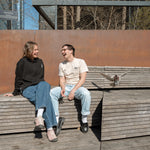 Two people sitting at the Spur benches, one wearing the Dinosaur Hoodie, the other wearing the Pigeon Pocket Tee—a pigeon lands next to them.