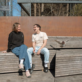 Two people sitting at the Spur benches, one wearing the Dinosaur Hoodie, the other wearing the Pigeon Pocket Tee—a pigeon lands next to them.