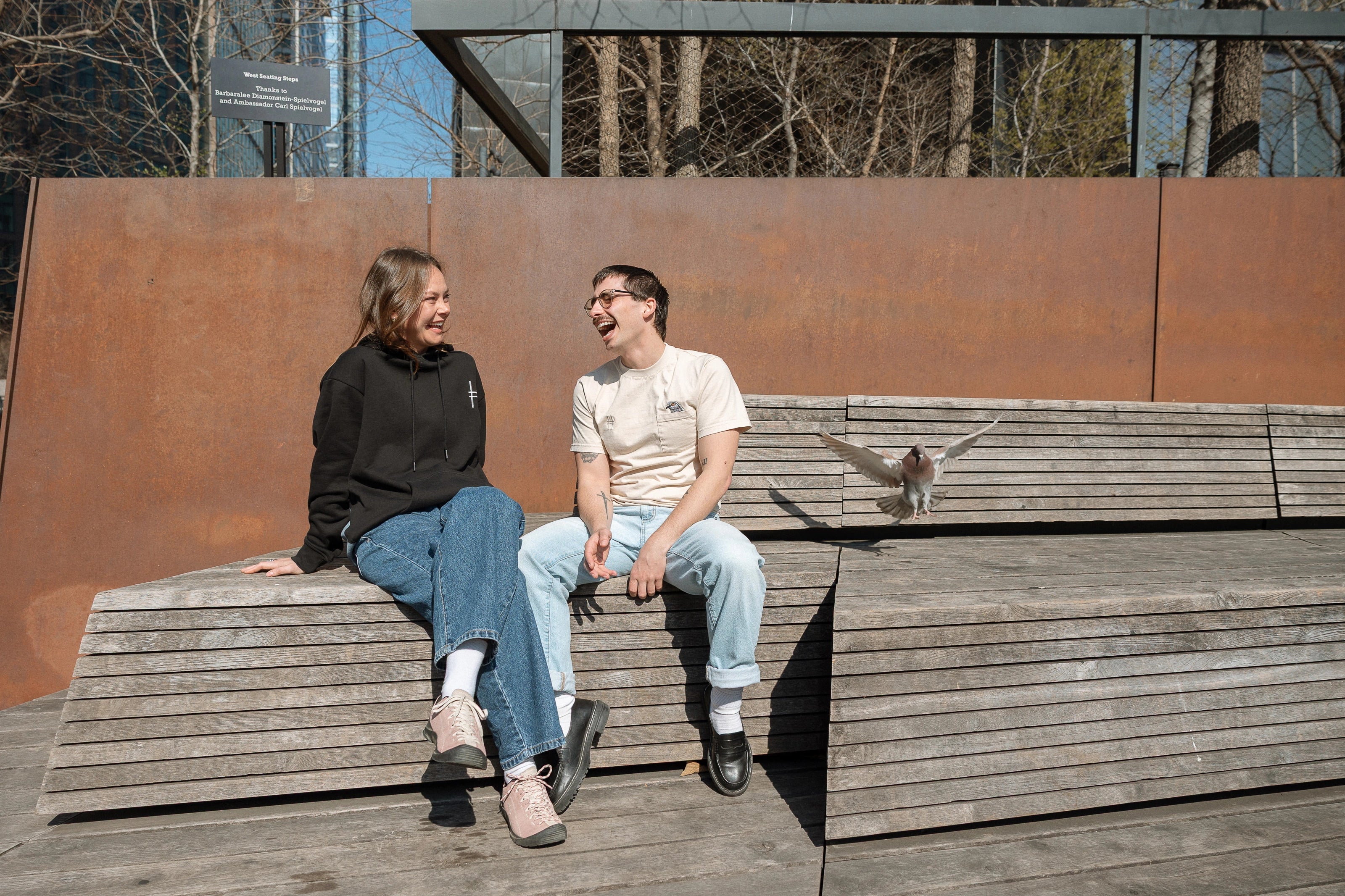 Two people sitting at the Spur benches, one wearing the Dinosaur Hoodie, the other wearing the Pigeon Pocket Tee—a pigeon lands next to them.