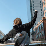 Woman wearing the Dinosaur Hoodie, arms outstretched against the blue sky and view down 10th avenue from the High Line northern spur.