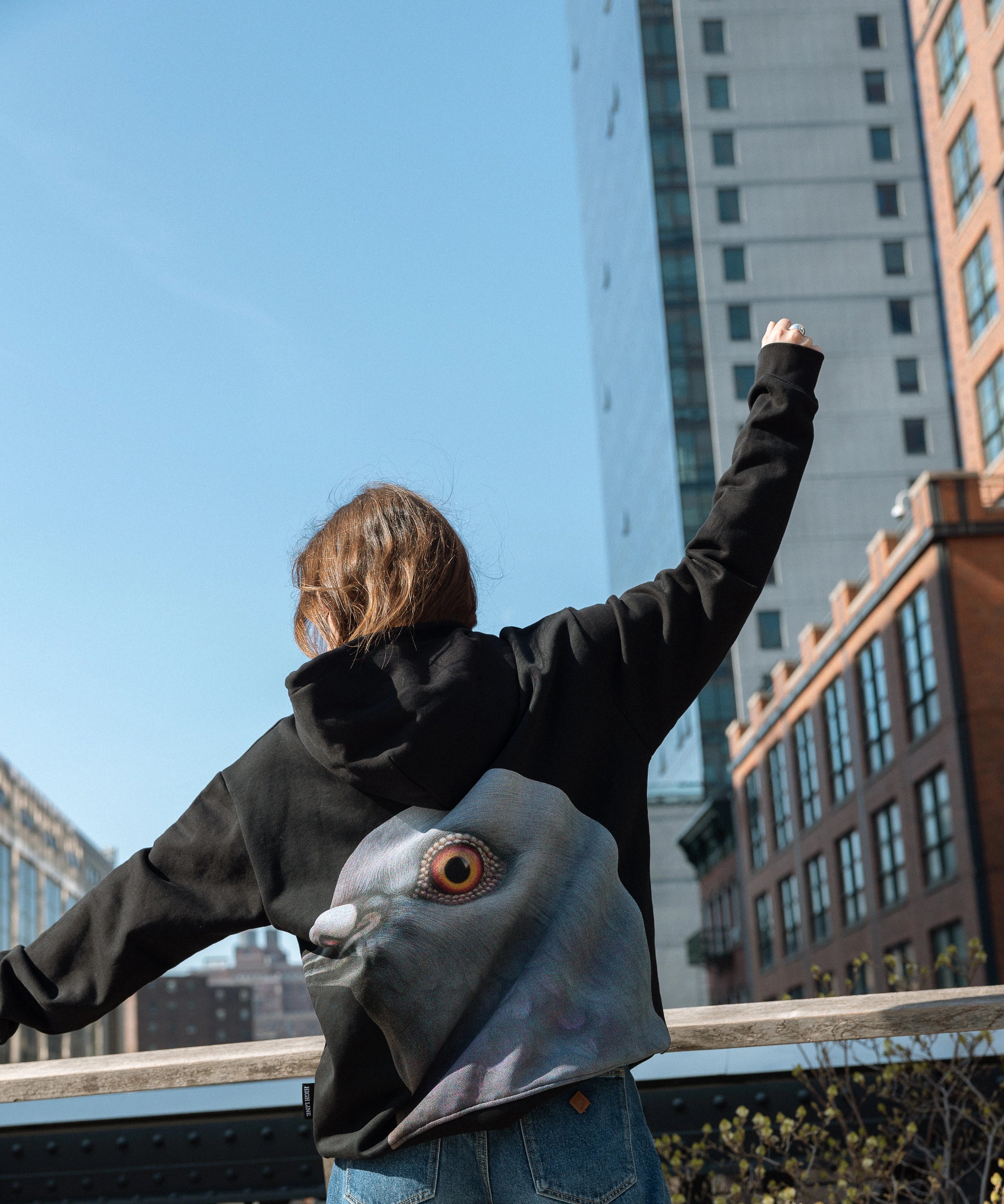 Woman wearing the Dinosaur Hoodie, arms outstretched against the blue sky and view down 10th avenue from the High Line northern spur.