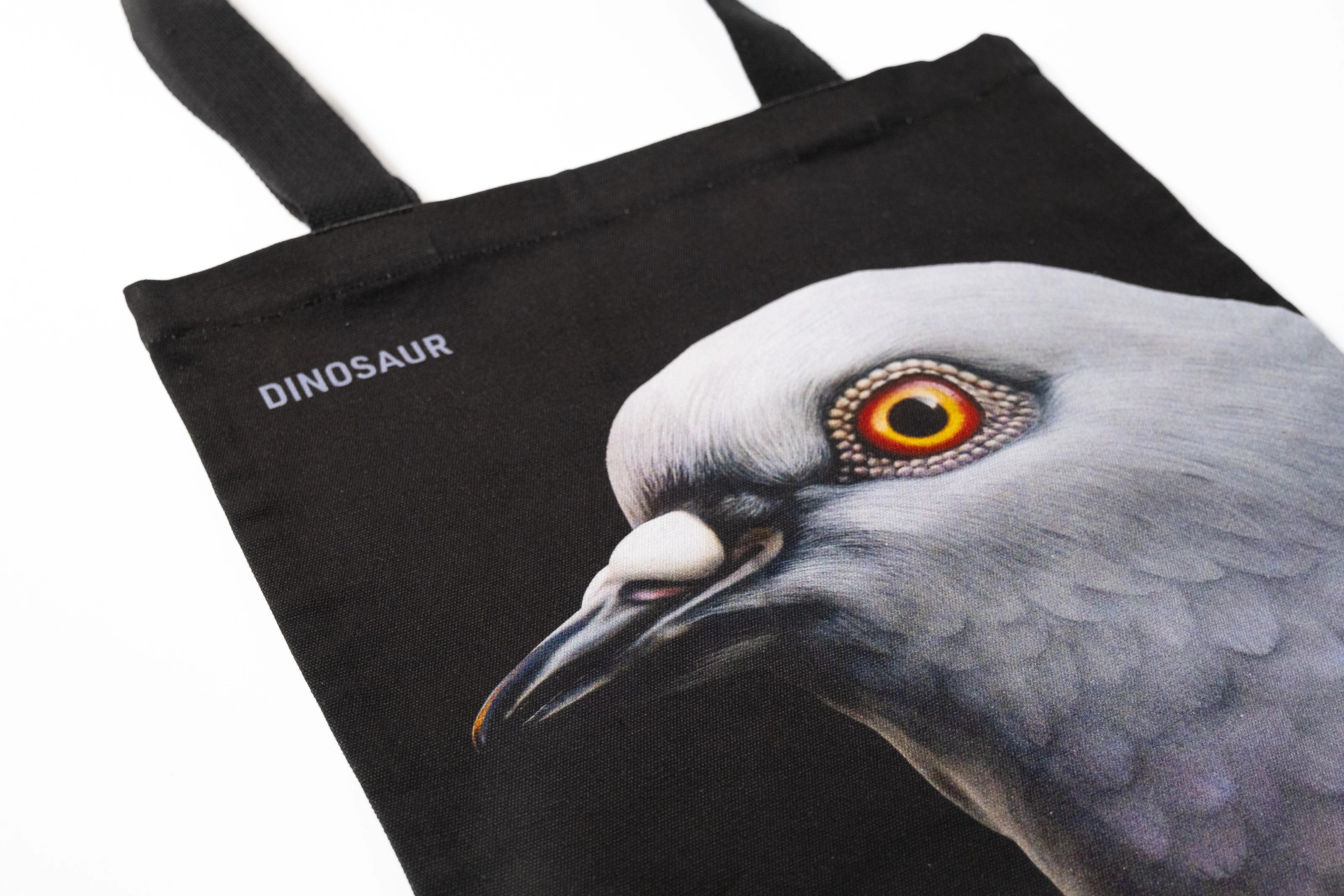 Closeup shot showing Dinosaur Tote front, with big pigeon head and "Dinosaur" screen-printed  on a black tote.