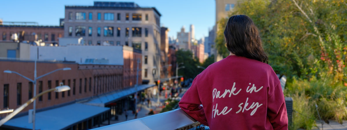 Woman leans on the railing as she looks south on the High Line. She wears the Park in the Sky Crewneck in Smokebush, a dark red crewneck with "Park in the Sky" appliquéd across the back in white script.