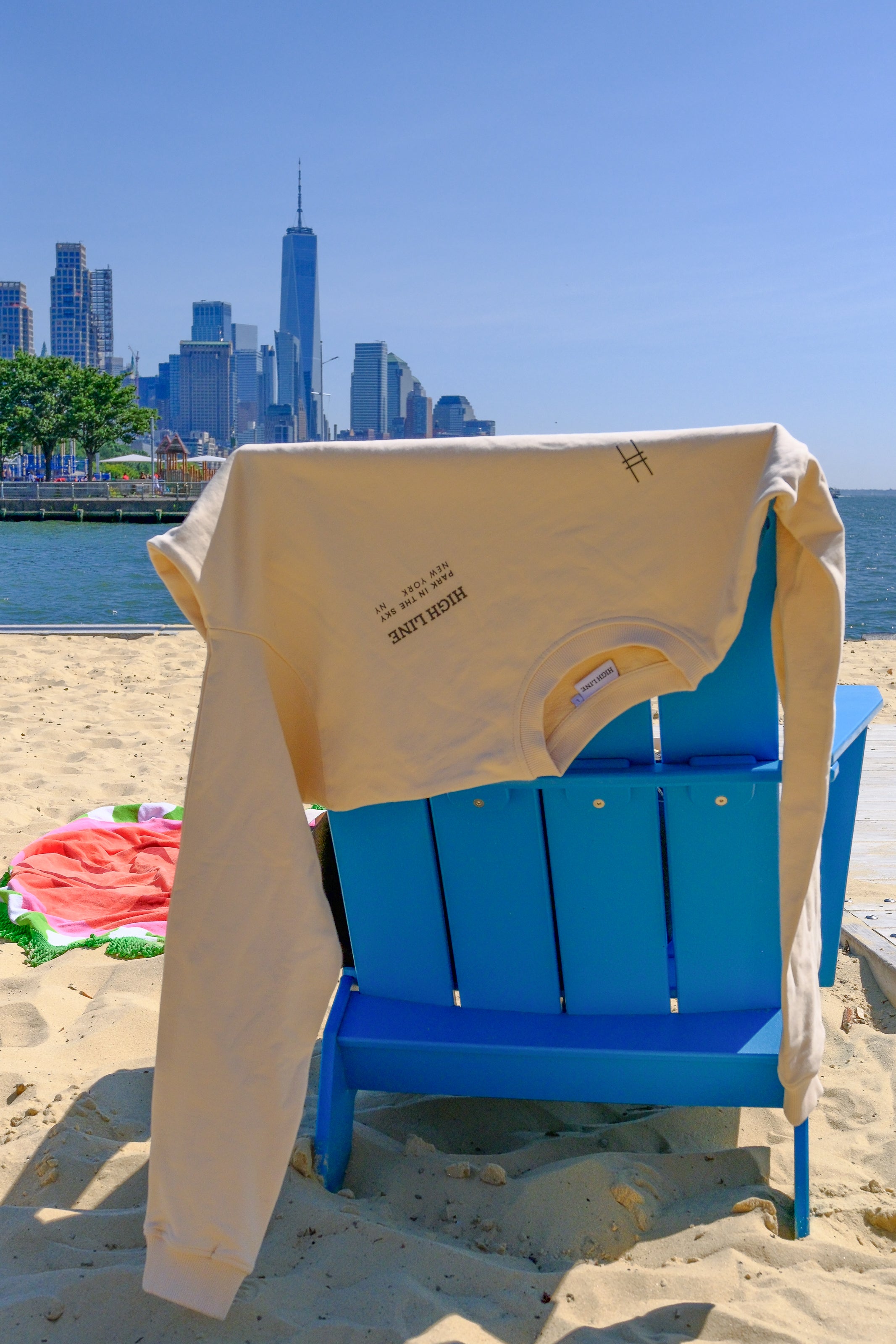 The High Line Crewneck Sweatshirt in River Birch hangs off the back of a beach chair on Gansevoort Beach with the NYC skyline in the background.