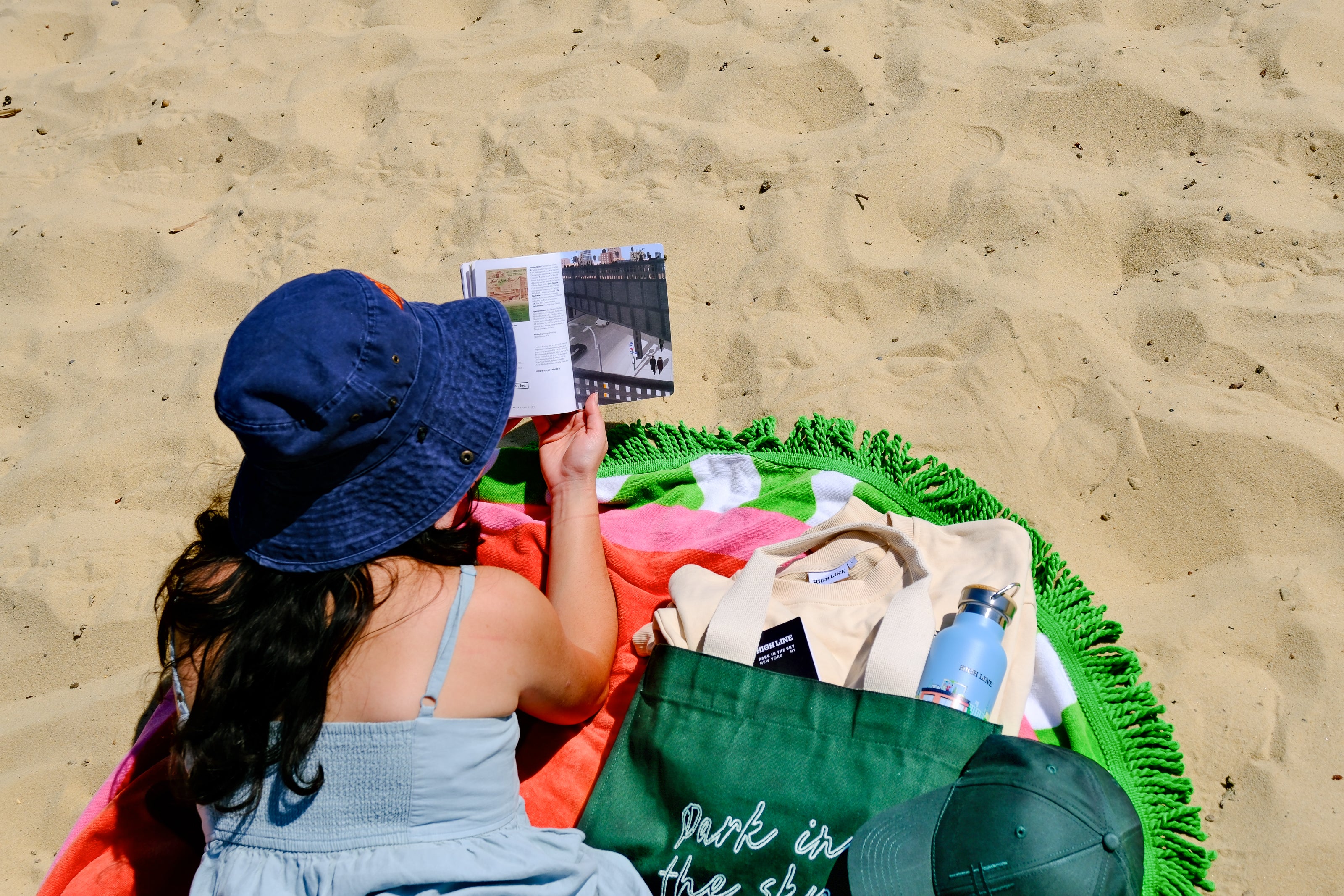 A person reclining on a sandy beach wearing a blue bucket hat and a light blue dress, holding a book with the NYC skyline and Hudson River in the background. She's reading the High Line Field Guide and has her water bottle, sweatshirt, and more in the Park in the Sky Tote in Ivy.