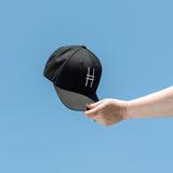 High Line Icon Contrast Cap—black cap with a white High Line icon embroidered on the front—held up against a clear blue sky.