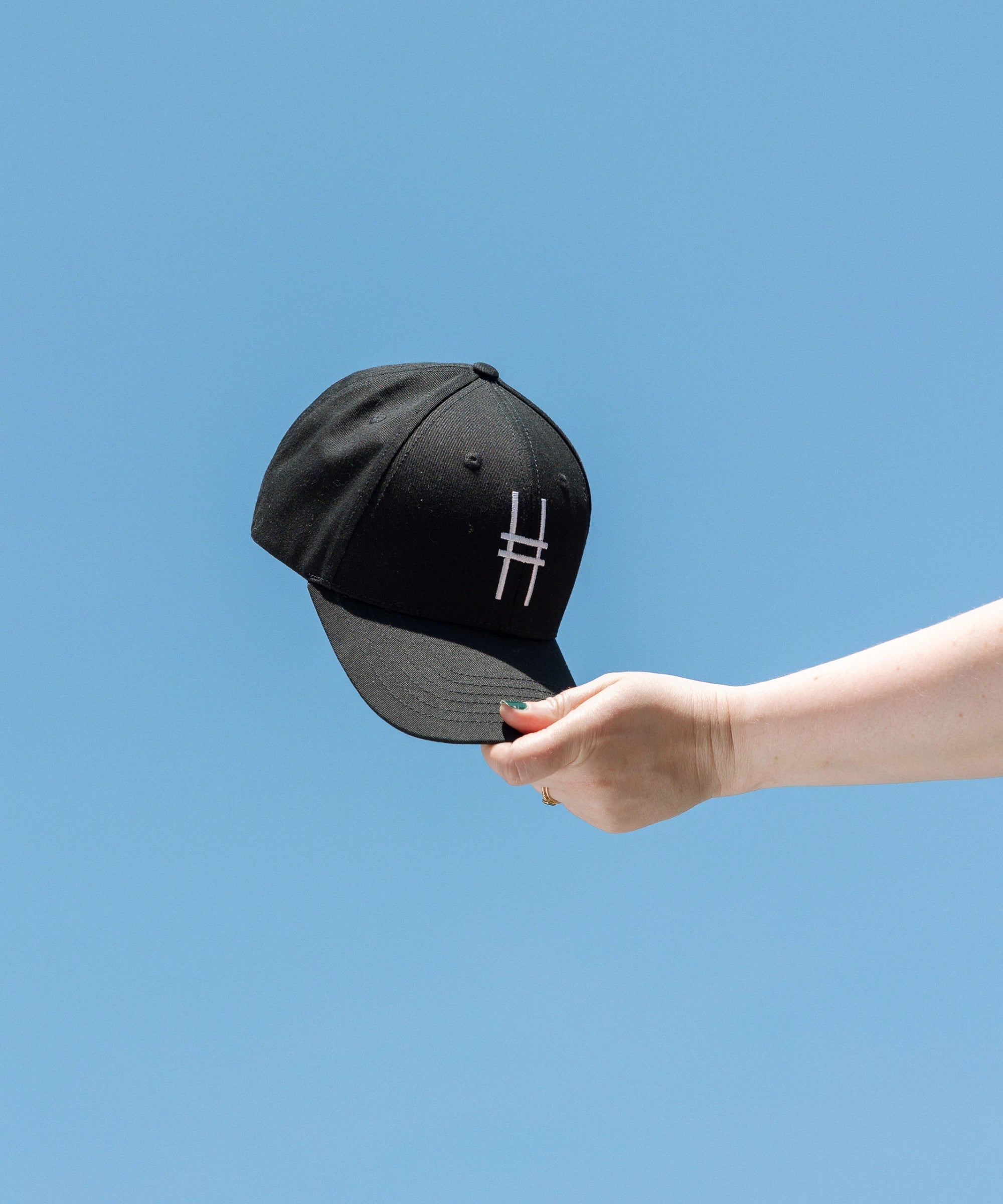 High Line Icon Contrast Cap—black cap with a white High Line icon embroidered on the front—held up against a clear blue sky.