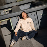 Woman wearing High Line Crewneck and sitting amidst the Pershing Square Beams on the High Line. Her eyes are closed as the sun shines on her.