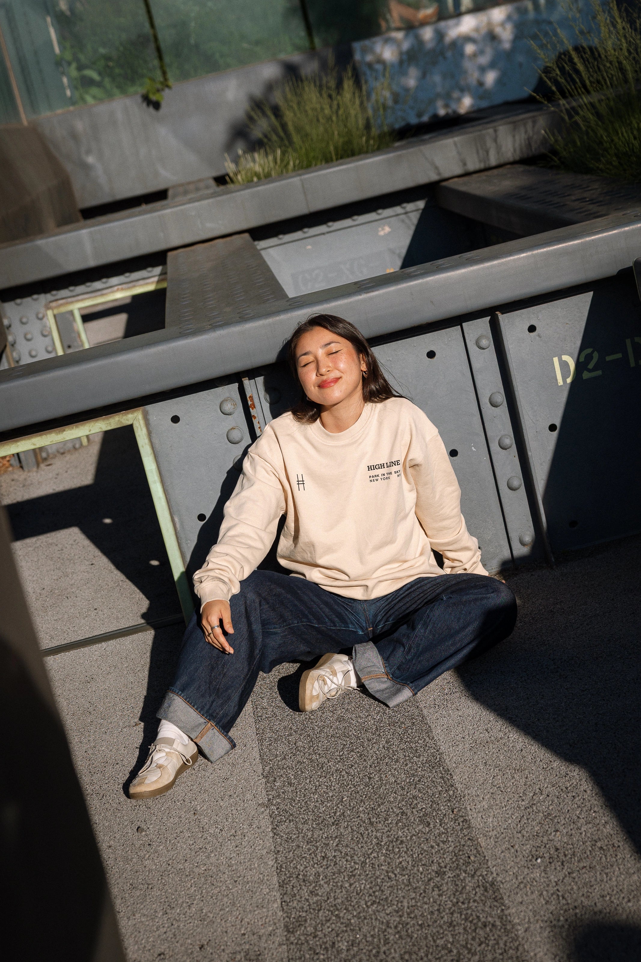 Woman wearing High Line Crewneck and sitting amidst the Pershing Square Beams on the High Line. Her eyes are closed as the sun shines on her.