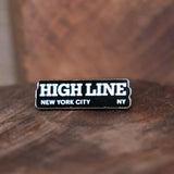 High Line Enamel Pin sitting on a dark wood block.