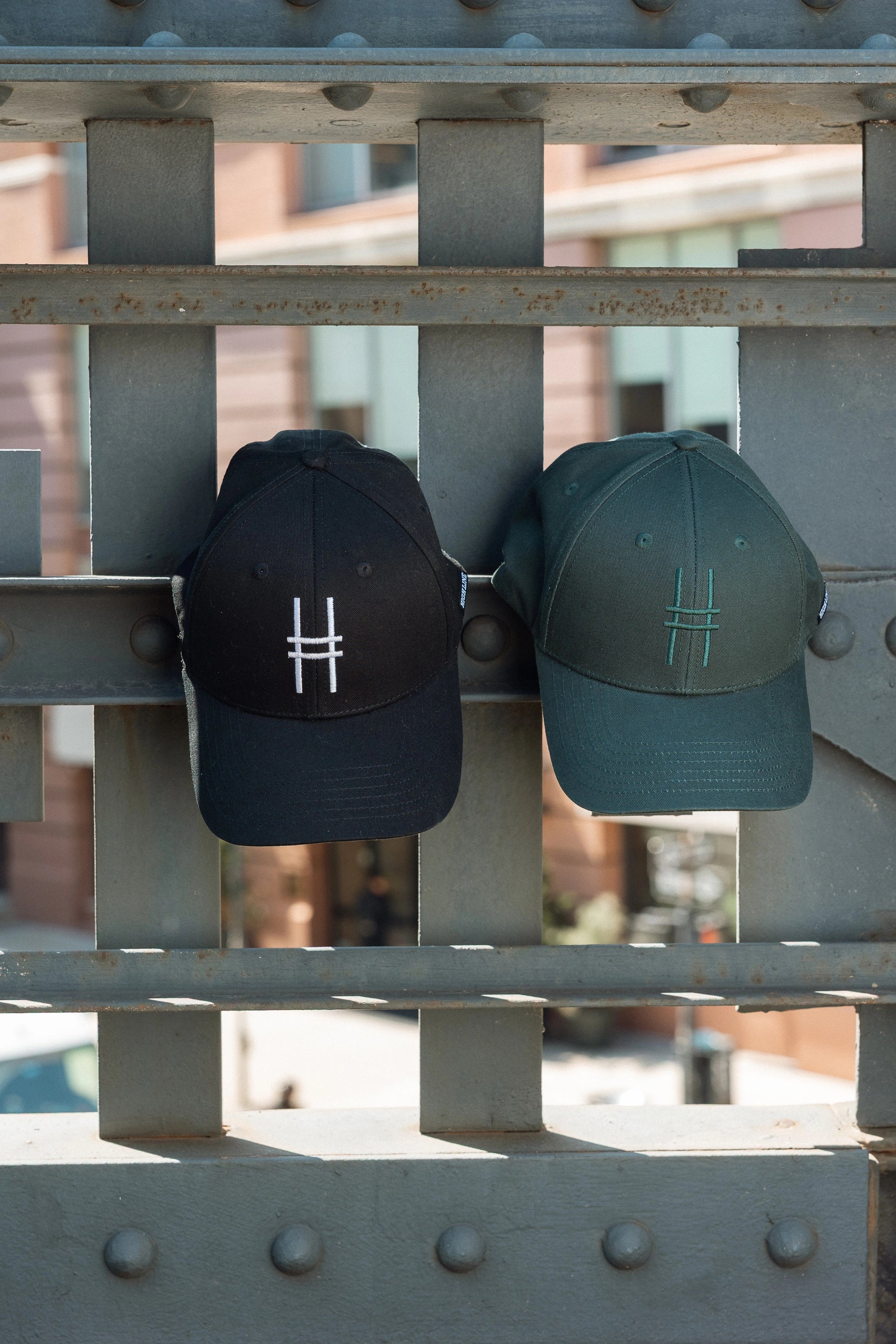 Both High Line Icon Caps side by side hanging off of the historic High Line railing.