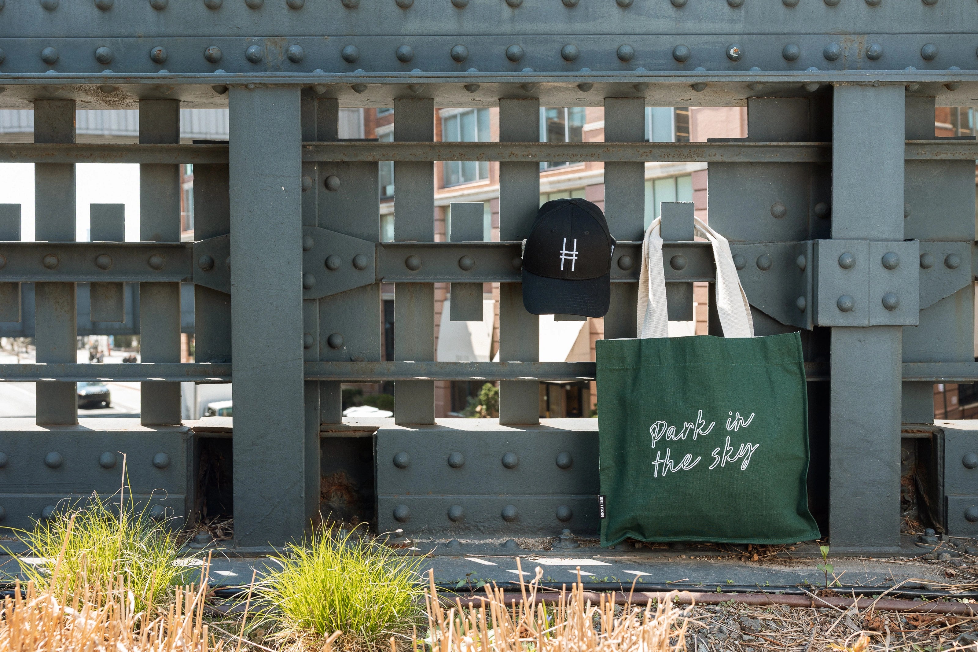 Park in the Sky Tote in Ivy and the High Line Icon Contrast Cap, hanging off of the railing of the High Line.