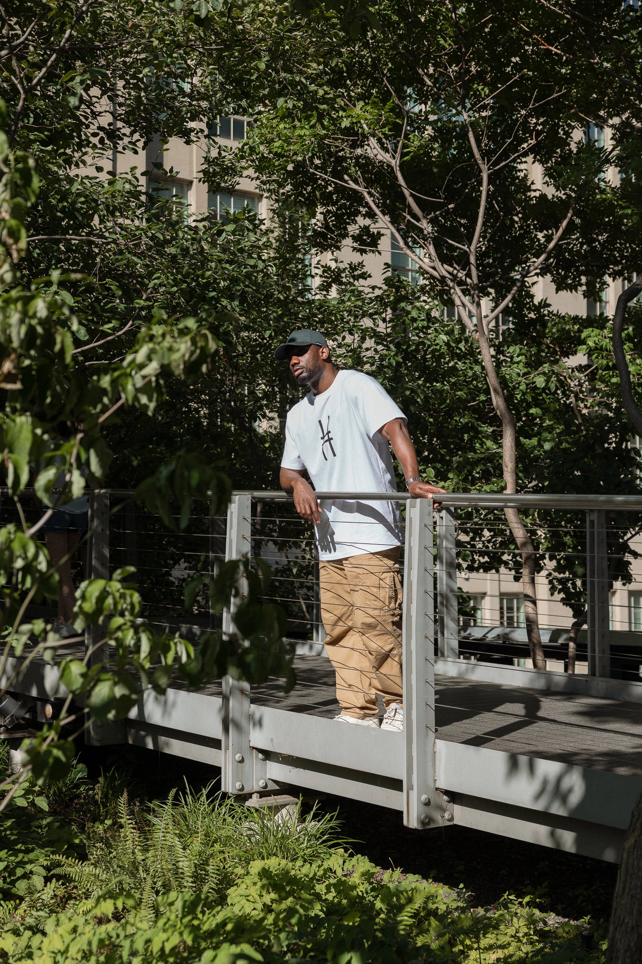 Man wearing the Big Icon Tee in white and High Line Icon Cap standing amidst the trees on the High Line.