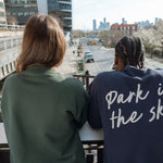 Two people looking down 10th Ave from the High Line, one in the Big Icon Crewneck in Ivy, one in the Park in the Sky Crewneck in Navy