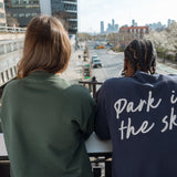 Two people looking down 10th Ave from the High Line, one in the Big Icon Crewneck in Ivy, one in the Park in the Sky Crewneck in Navy