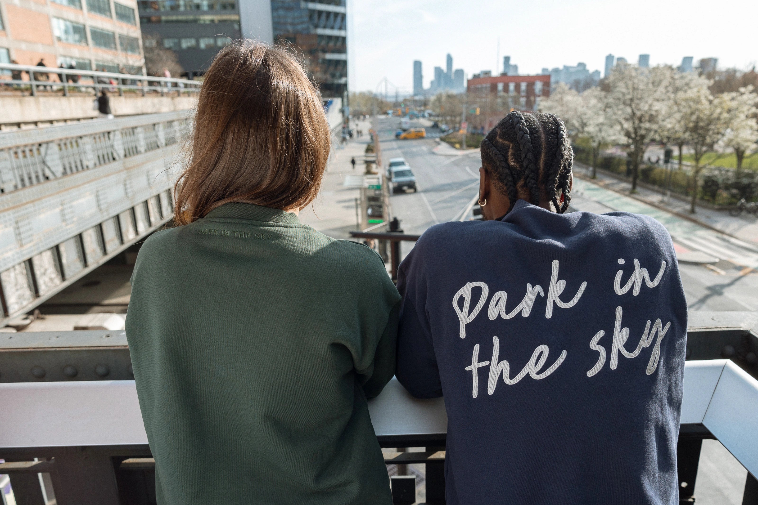 Two people looking down 10th Ave from the High Line, one in the Big Icon Crewneck in Ivy, one in the Park in the Sky Crewneck in Navy